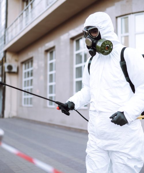 Man in protective suit and mask sprays disinfector onto the railing in the public place. Covid -19.