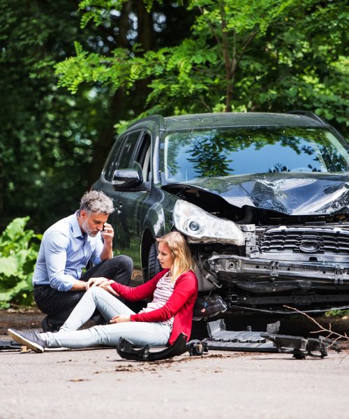 Young woman by the car after an accident and a man making a phone call.
